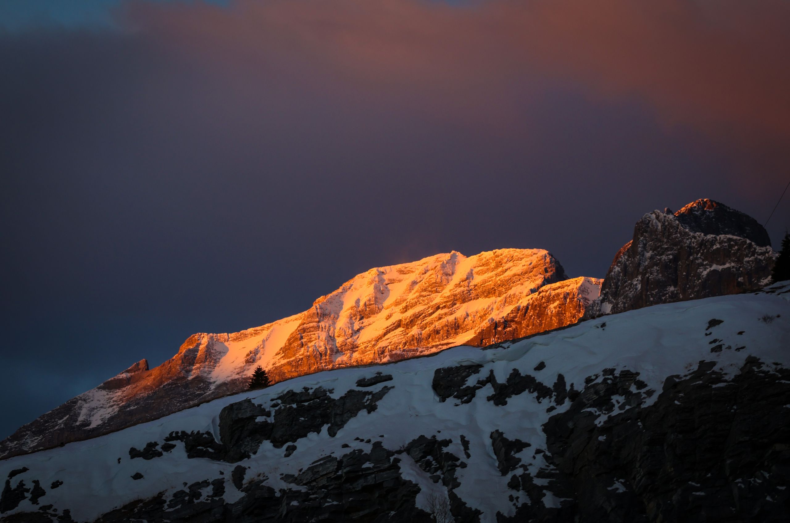A setting or rising sun bathes the summit of one of the Rocky Mountains in golden light, while the rest of the range sits in relative darkness.