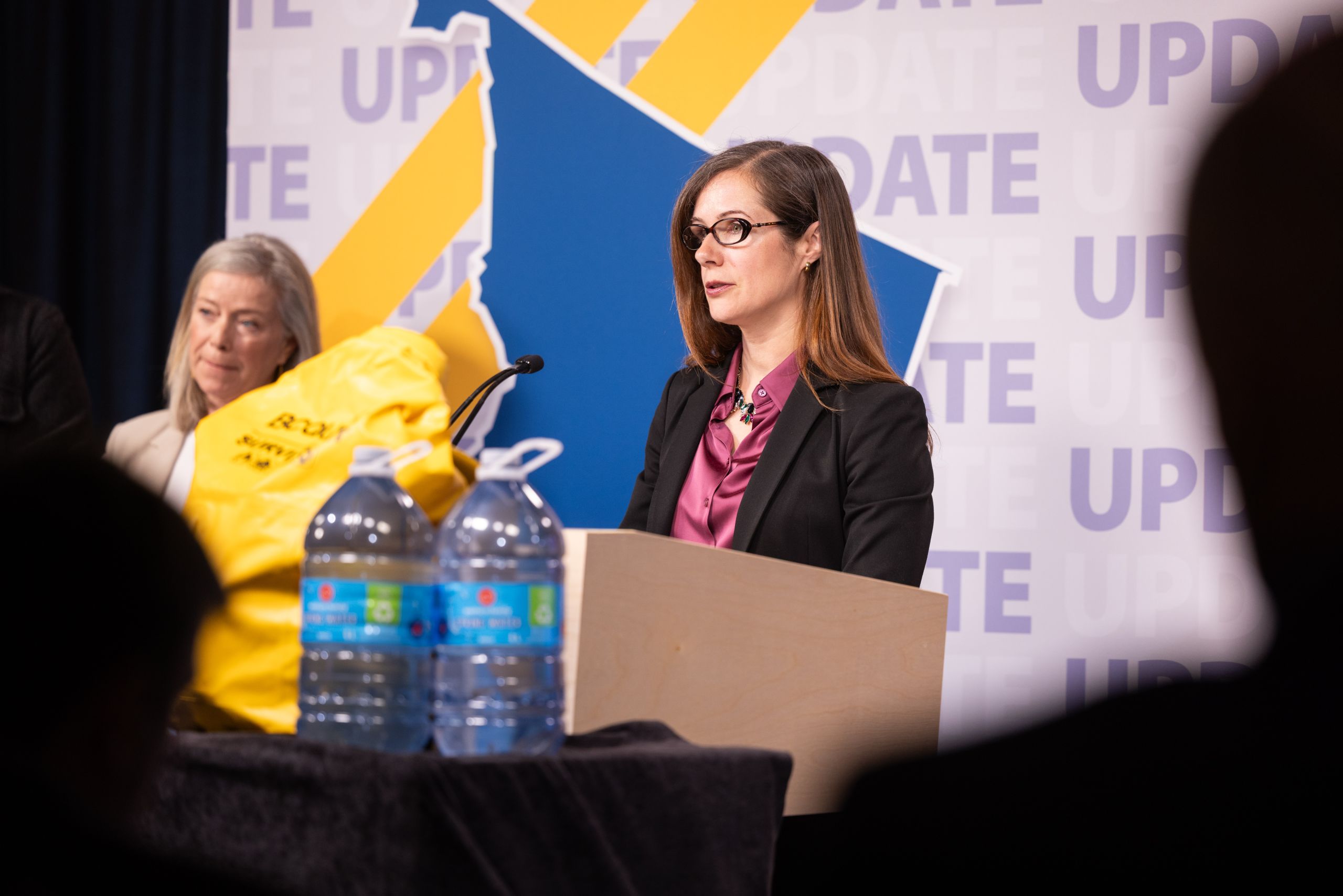 BC Emergency Management and Climate Readiness stands at a podium with a blue and yellow design behind her. There are two bottles of water in the foreground