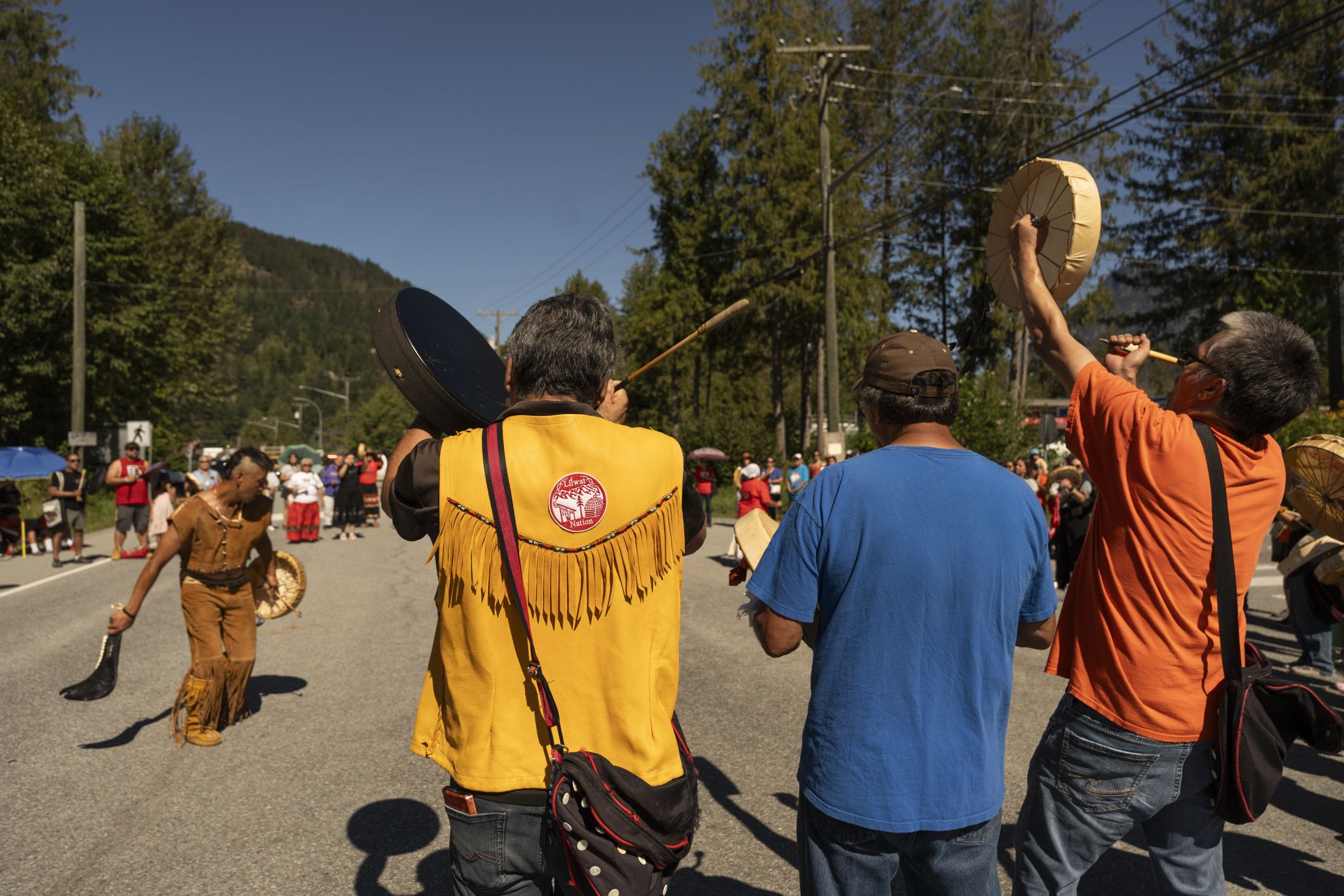 On Lil'wat territory in Mount Currie, a crowd stands in the middle of the road. Many wear regalia and hold drums. To the left, in the middle of the crowd, a dancer wearing buckskin regalia makes is way dancing in the circle. The sun is bright, the sky is stark blue