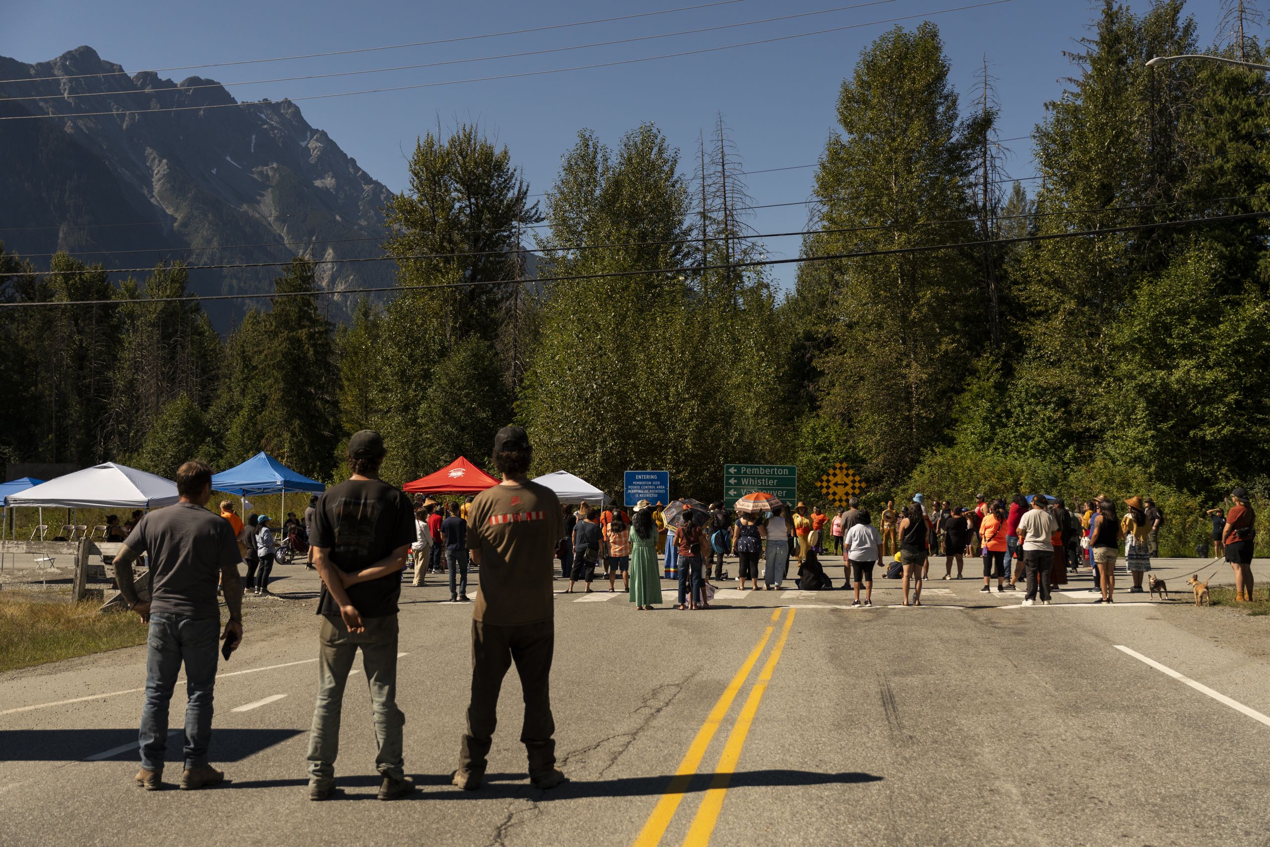 Three men stand back from the crowd on Highway 99 at Mount Currie while Líl̓wat and N'Quatqua nations hold ceremony and protest B.C. opening Joffre Lakes Park.