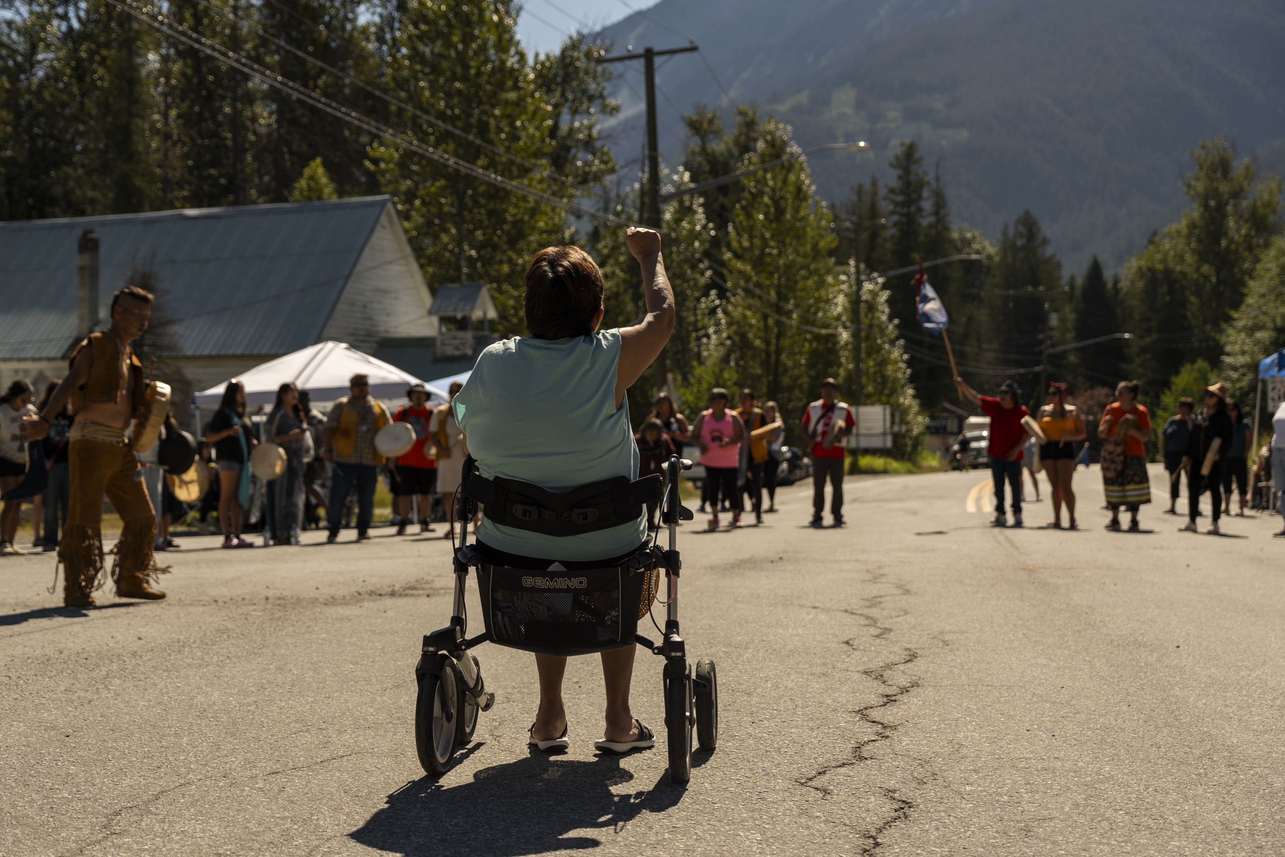 At a Lil'wat road closure protesting B.C. keeping Joffre Lakes open, a woman sits in a walker facing away from the camera towards a crowd standing in a circle on the road. She holds her fist up in solidarity. Mountains are visible in the background.