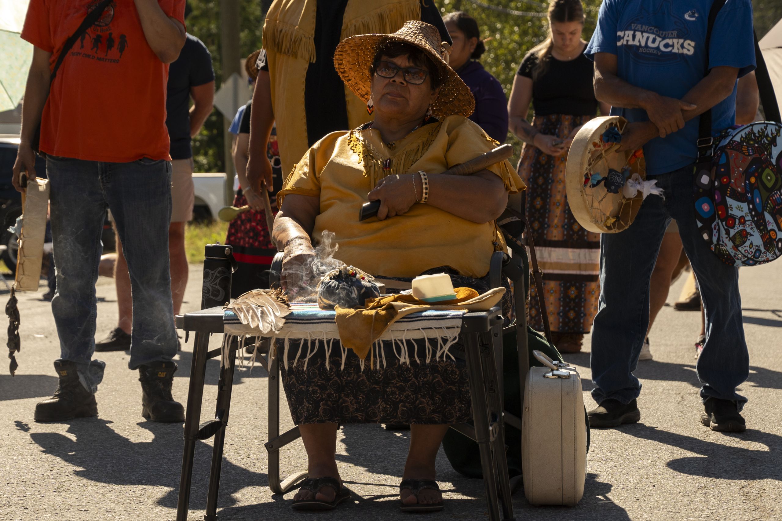 In Mount Currie where Líl̓wat and N’Quatqua Nations held a ceremony and road closure, Linda Dan sits in the sun, wearing a cedar hat, with a ceremony table in front of her. She wears sunglasses and is surrounded by supporters