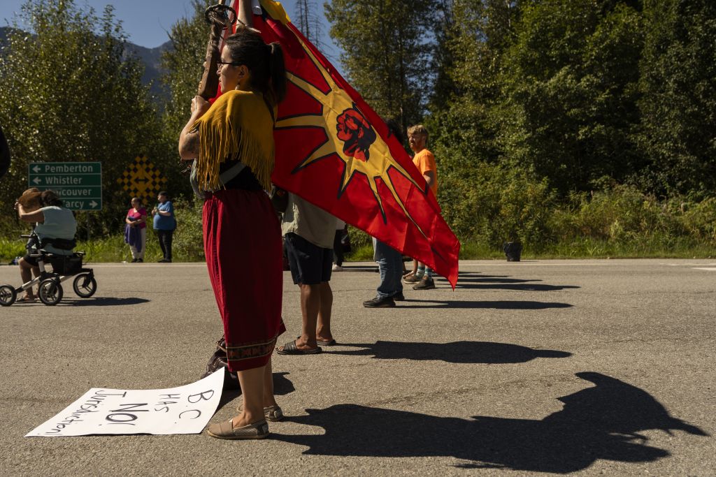 Amanda Ritchie stands in profile, facing left. She wears red and holds a red flag known as a unity flag, which depicts a long-haired Indigenous man in profile against a bring yellow sunburst