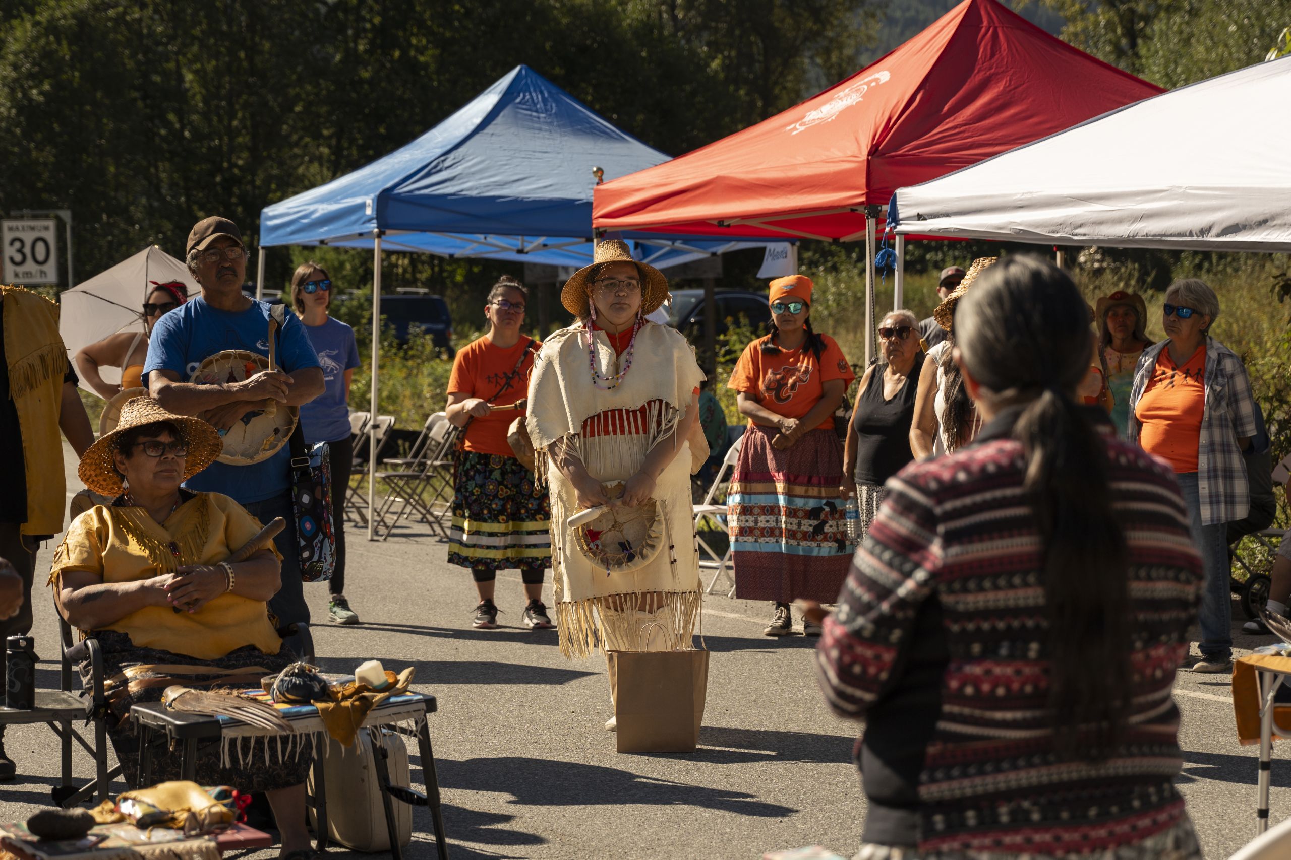In Mount Currie, Casey Gonzalez, director of k̓wezúsmin̓ (title and rights) for Líl̓wat, stands in light buckskin regalia and a cedar hat holding a drum and listening to people speaking in the centre of the crowd at the road closure led by Líl̓wat and N’Quatqua Nations.