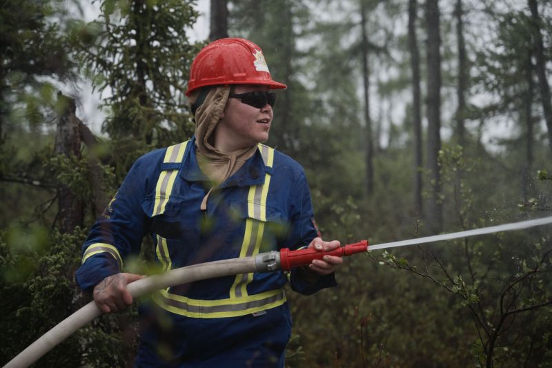 A woman in blue safety overalls and a hard hat holds a firefighting hose and sprays water toward the right.