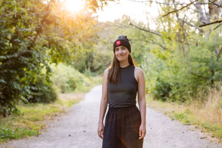 One of The Narwhal's new assistant editors, Paloma Pacheco, is seen standing on a trail through a greenspace wearing a Narwhal touque. The sun sets over her right shoulder.