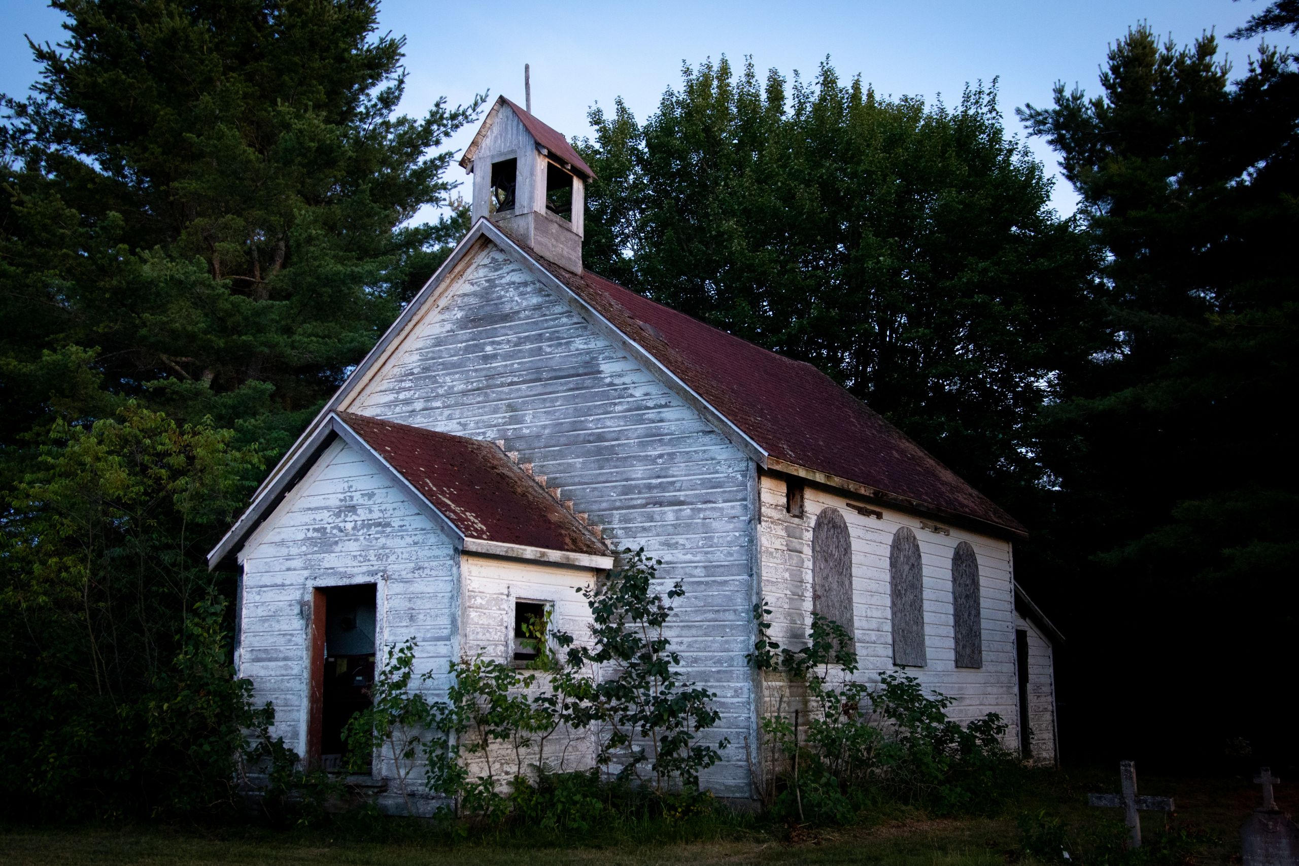 An abandoned church on the Shawanaga First Nation, which is home to hundreds of bats, with trees behind it and overgrown bushes in front.