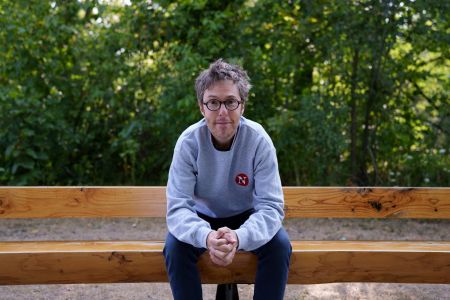 Will Pearson, a new Narwhal assistant editor, sitting on a bench in Peterborough, Ont. with greenery behind him. He is wearing a grey sweatshirt with a red Narwhal logo and clasping his hands together.