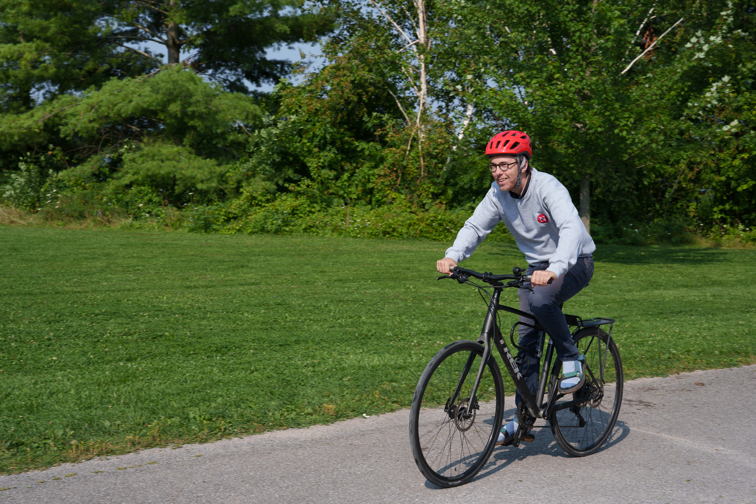 Will Pearson riding a bike on a sunny day in a green park. He's wearing a grey Narwhal sweatshirt and red helmet.