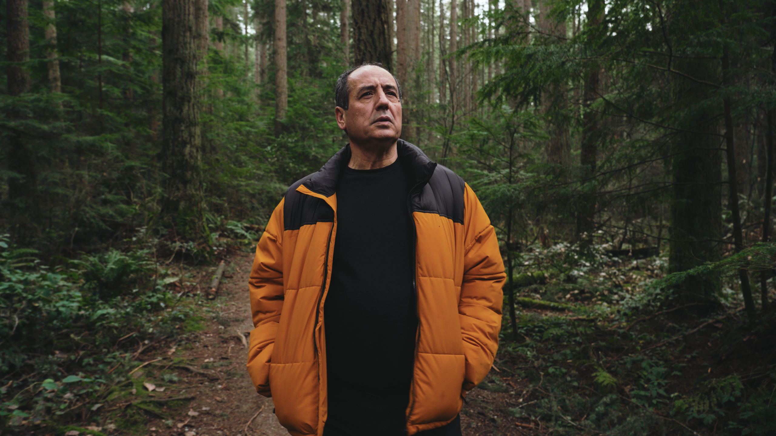 A portrait of Younes Alila wearing a yellow and black coat in the forest