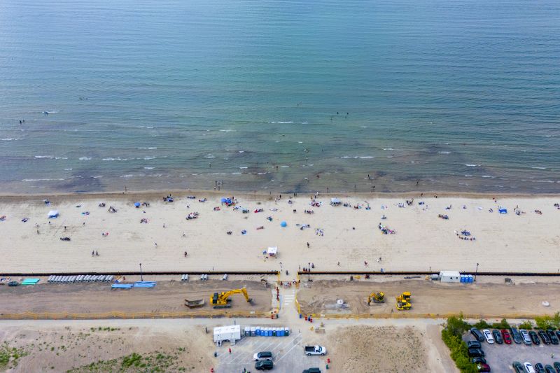 An overhead drone photo of Wasaga Beach. On the bottom: a parking lot half full of cars and bulldozers digging a roadway through a construction zone. In the middle: bathers on the sandy beach. On the top: a turquoise Georgian Bay.