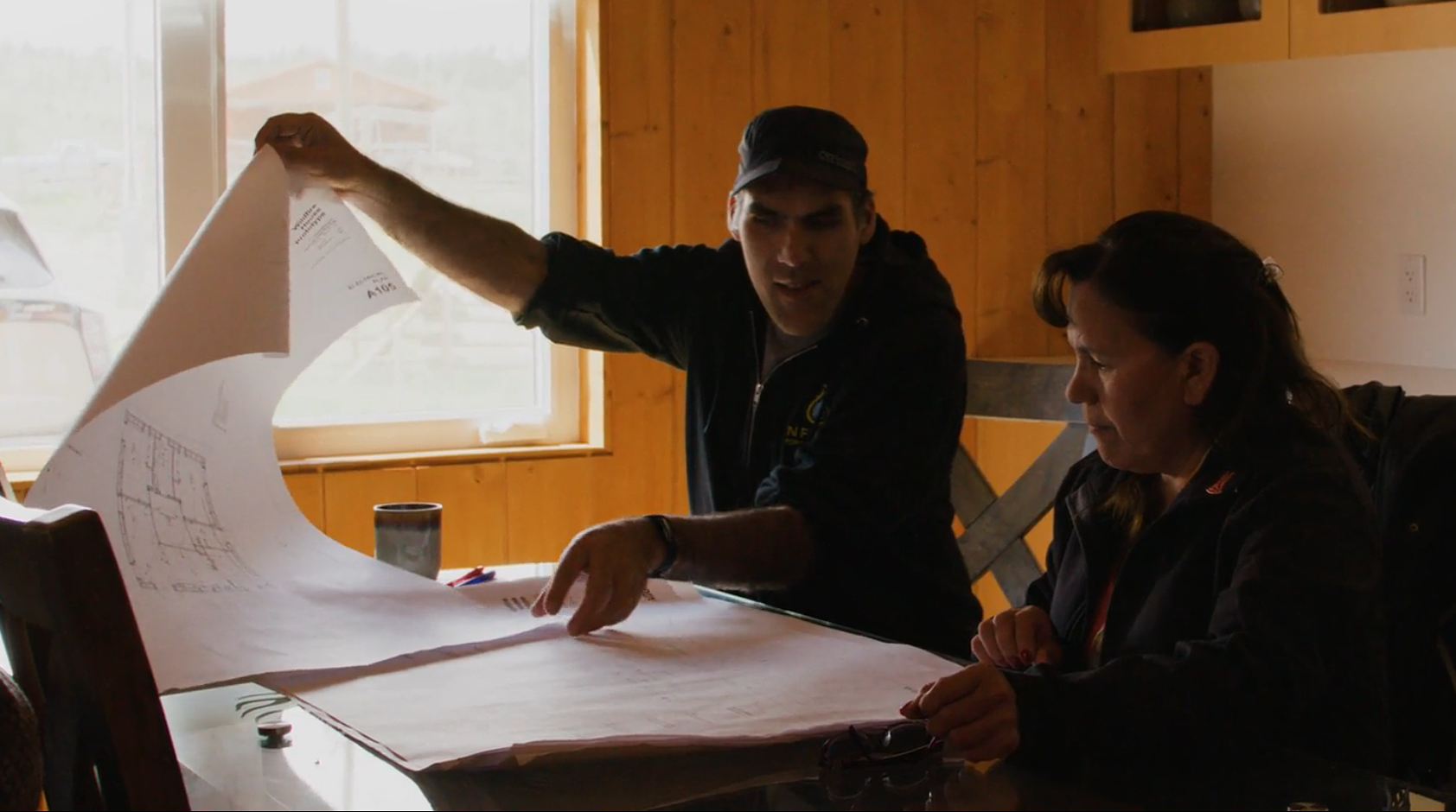 Russell Myers Ross sits at a table with a woman and spreads out large blueprints of the wildfire house design