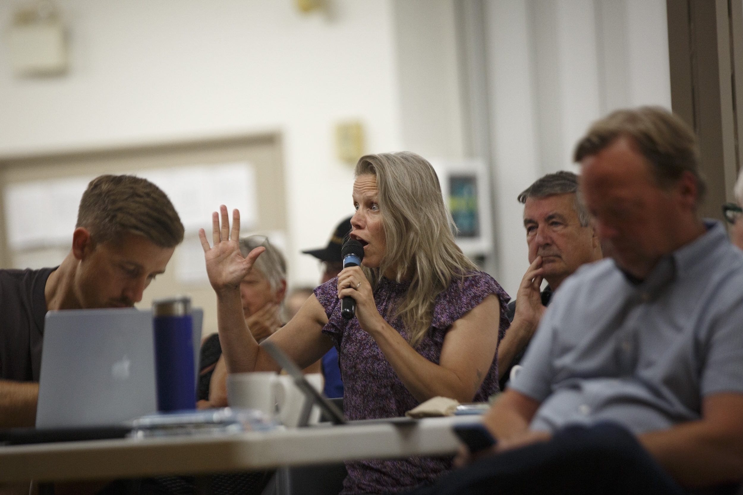 A woman with an animated expression and a raised hand speaks at a public meeting