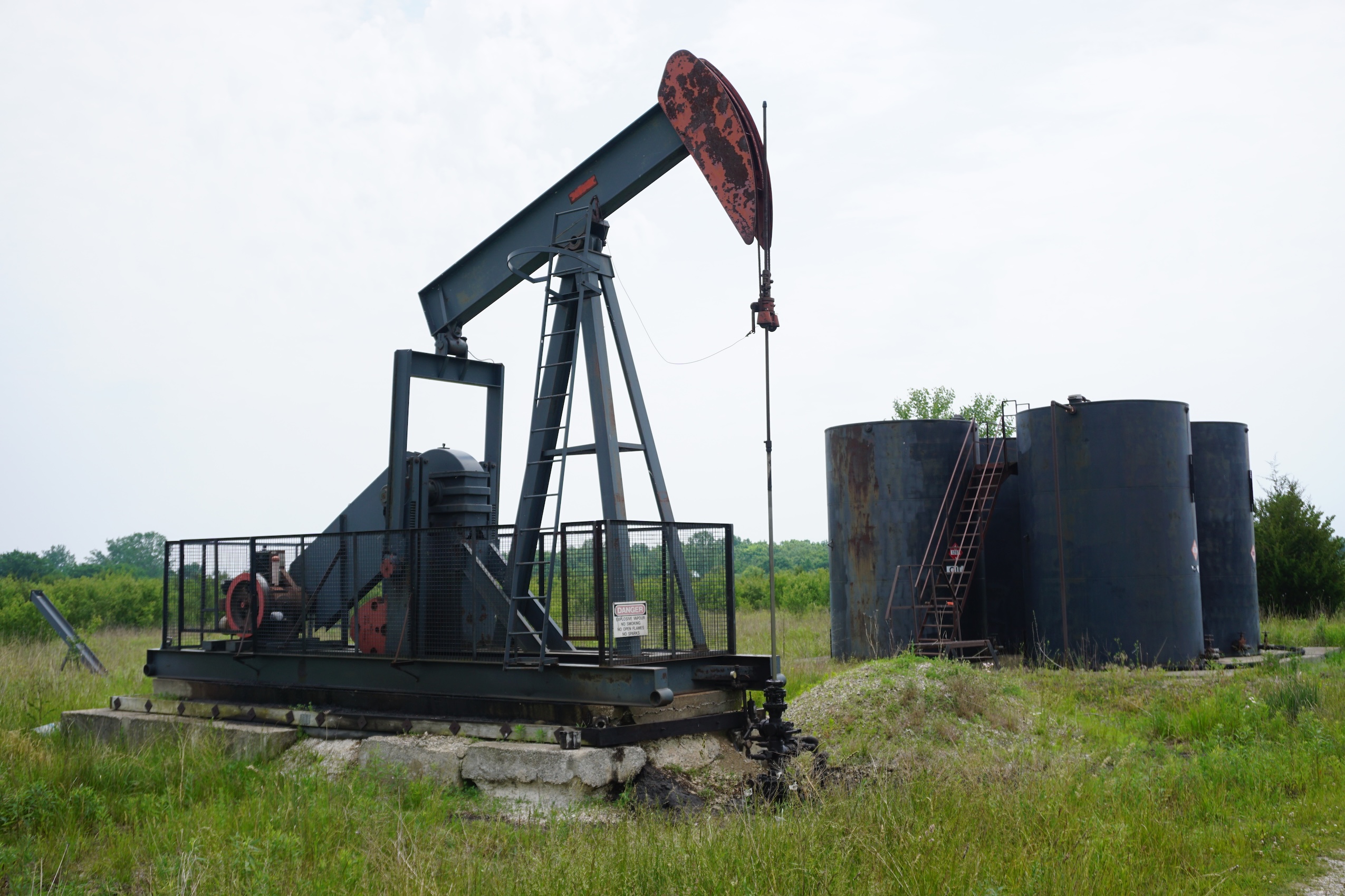 A black oil well is seen in the foreground, and four black storage tanks in the background.