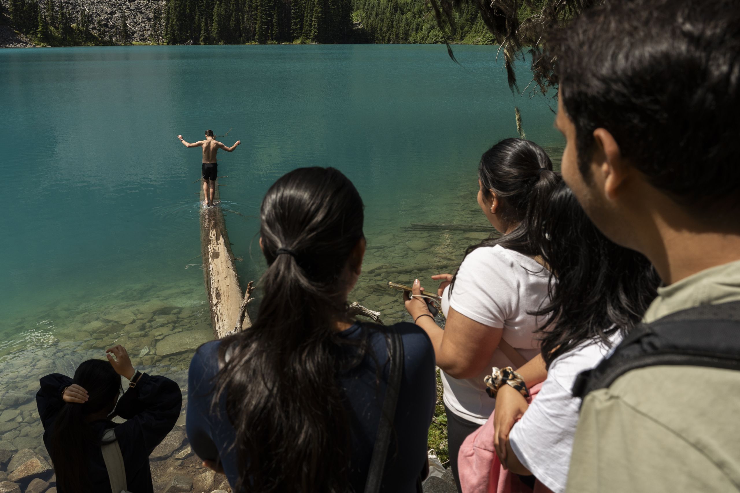 The camera view is from among the crowd around the famous log at the second lake at Joffre Lakes, while one person walks out onto the lake in swim shorts with their arms out for balance.