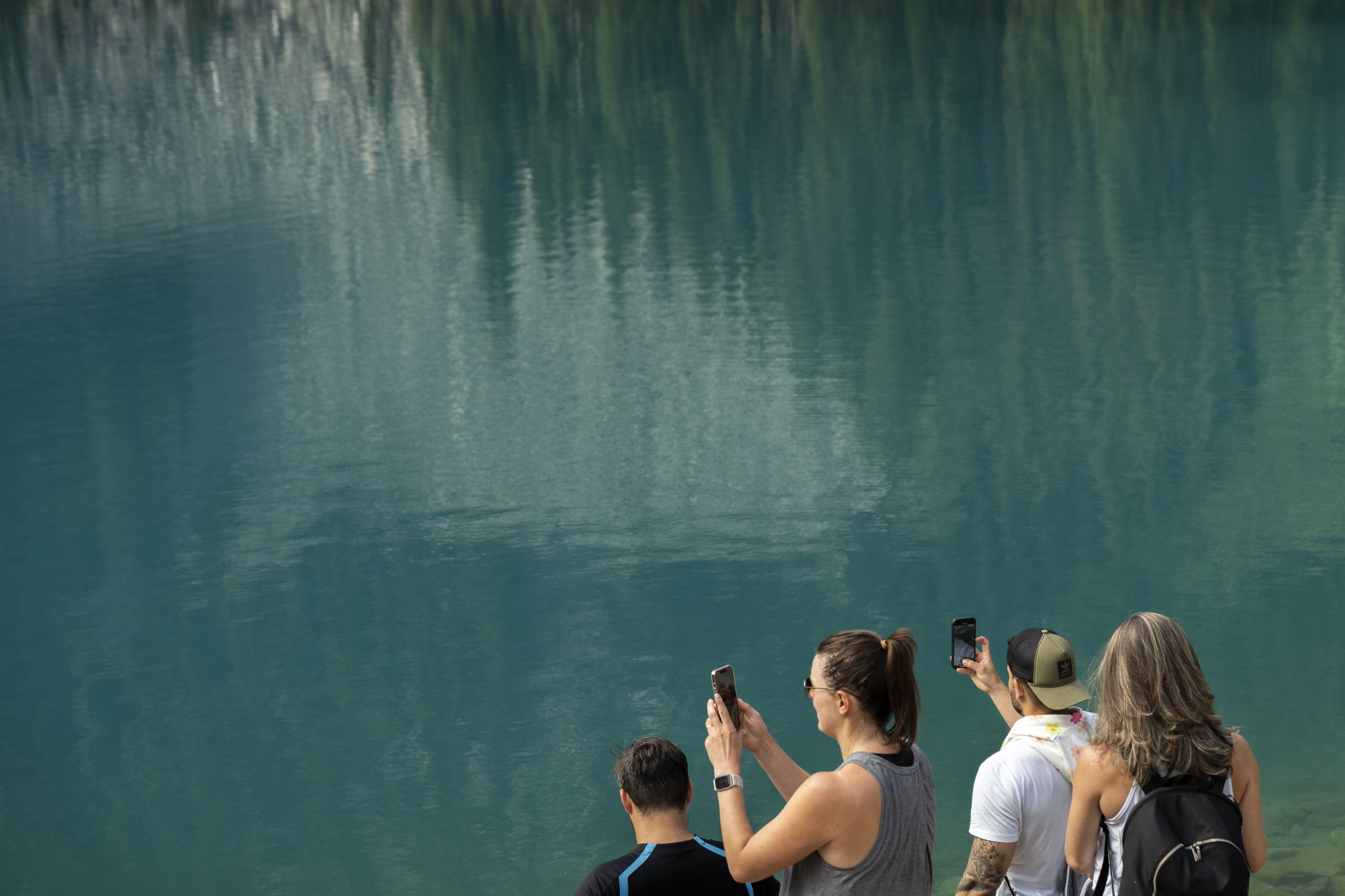 Most of the photo is taken up by the turquoise blue water at Middle Joffre Lake. In the bottom right corner, a group of hikers face left, holding up their phones towards the water.