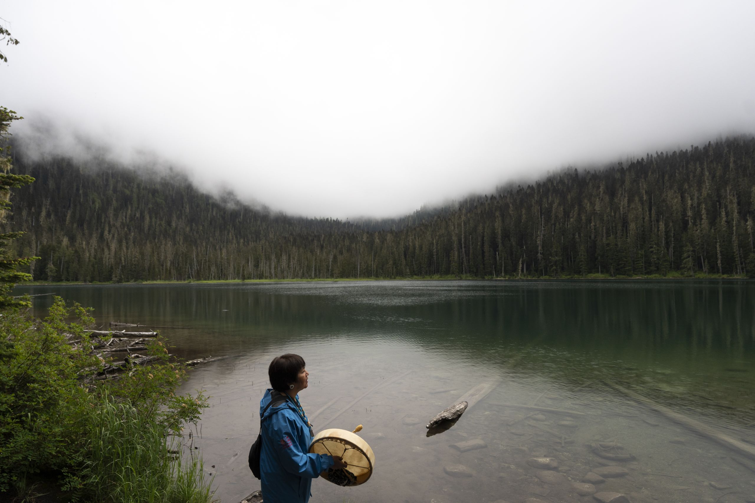 At Joffre Lakes, Maxine Bruce stands against the calm first lake, holding her drum, facing to the right in profile. Clouds hang low over the trees. She wears a blue jacket and sings.