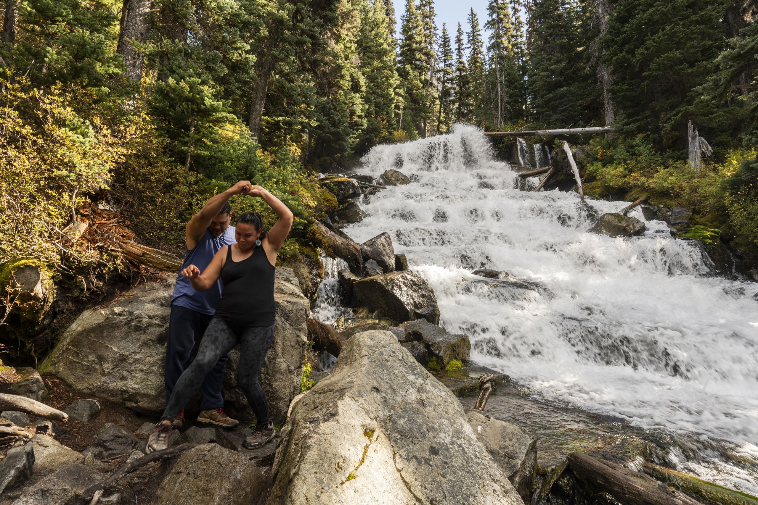Lil'wat community members Terry Jameson and Lightning Rose Jameson enjoy a peaceful moment walking along the waterfall on the trail at Joffre Lakes / Pipi7íyekw. He helps her walk down the rocks, but the ways their arms are raised as they hold hands, it almost looks like he is spinning her, dancing.