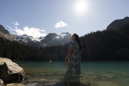 At Joffre Lakes, Lil'wat member Keisha Andrew stands in the green water under the bright sun and blue sky, looking upwards to the left, smiling. She wears a dress and is knee deep in the water. The peaceful moment happened during a recent reconnection period / closure at Joffre Lakes Park.