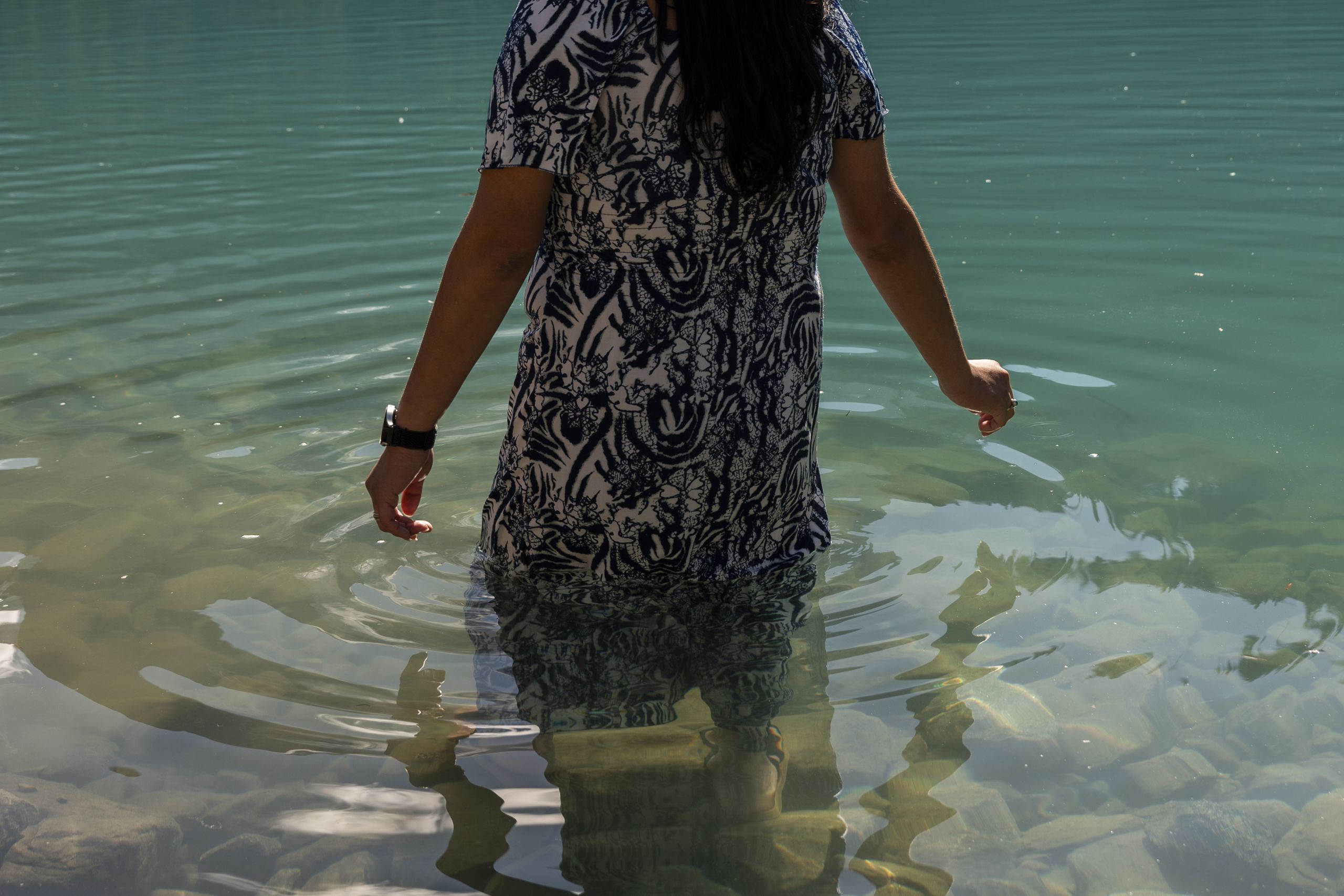 Keisha Andrew walks into the water at the middle lake at Pipi7íyekw during the re connection period in September. She wears a dress and is her fingers just graze the surface of the water.