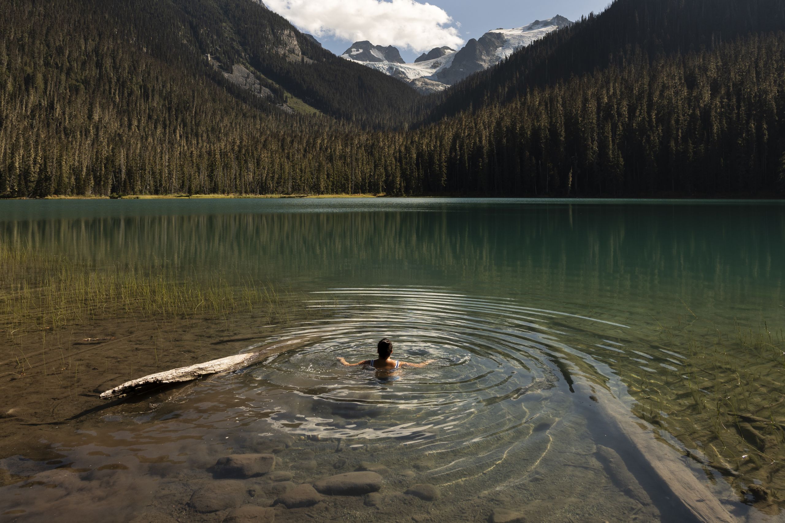A wide view of the middle Joffre Lake, where Jessie-Lynne Joe is swimming out from the shallow bank into the turquoise water. The lake is empty around her.