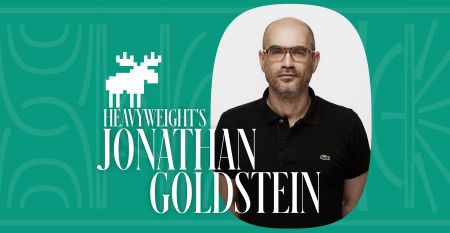 A photo of Heavyweight host Jonathan Goldstein, who is wearing a black Lacoste shirt and glasses. It is inside a teal background with a pixelated image of a Moose and a title reading "Heavyweight's Jonathan Goldstein."