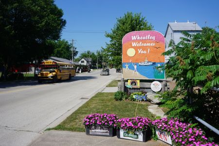 A colourful sign with a painted beach scene reads, "Wheatley welcomes you." It's on a residential street near a school bus and flower boxes