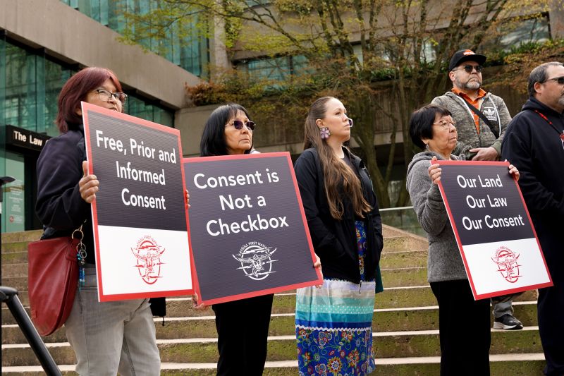 Xatsull community members hold signs that say "free, prior and informed consent" and "consent is not a checkbox" on the steps of the B.C. Courts