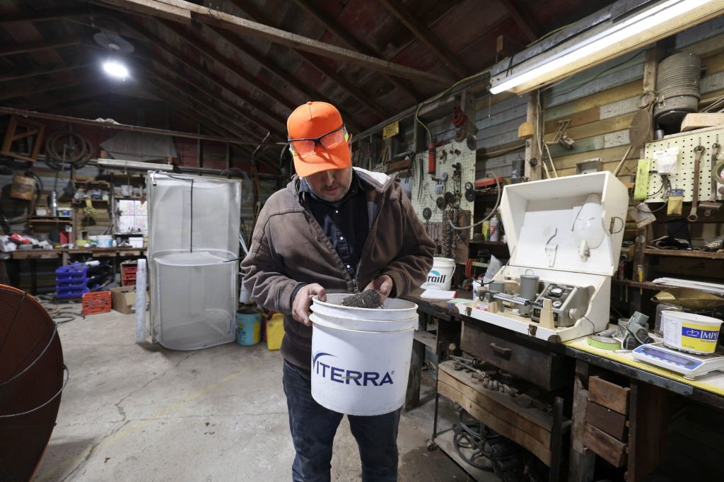 A man wearing an orange baseball hat sifts through a bucket of grains in a storage shed.