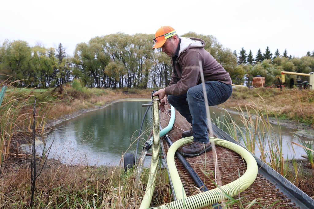 A man wearing an orange baseball hat handles a wide hose in front of a pond of water