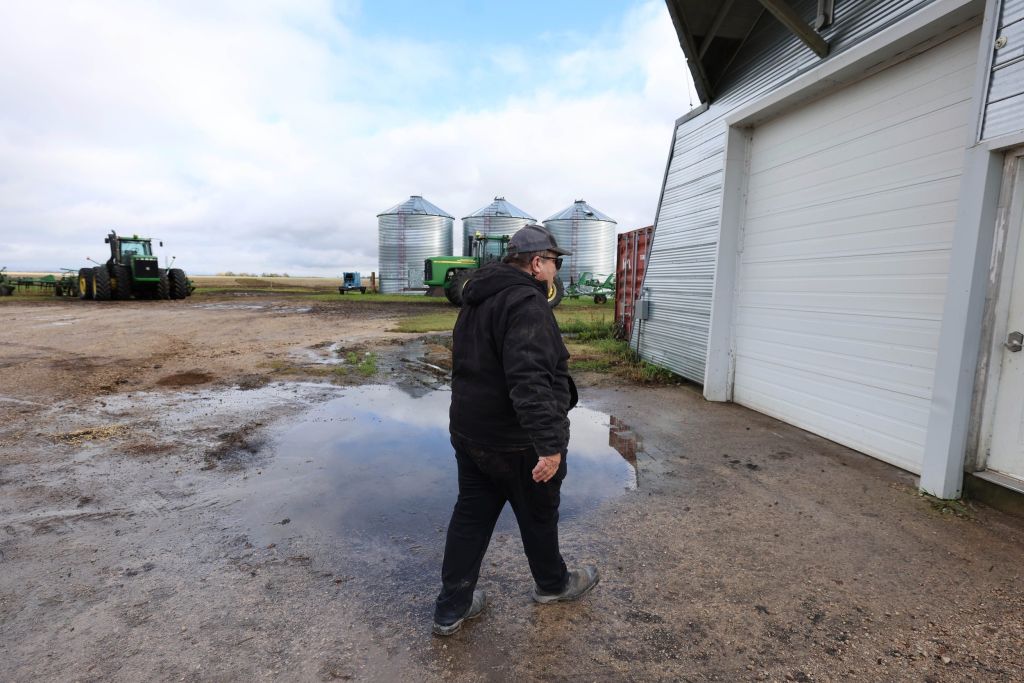 A man with his back to the camera walks toward a garage door, with large steel granary barrels in the distance.