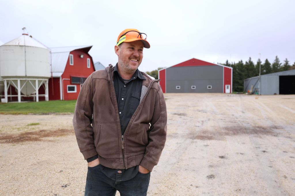 A man wearing a brown sweatshirt and orange baseball hat smiles and looks away from the camera. There are two red barnhouses in the background behind him.
