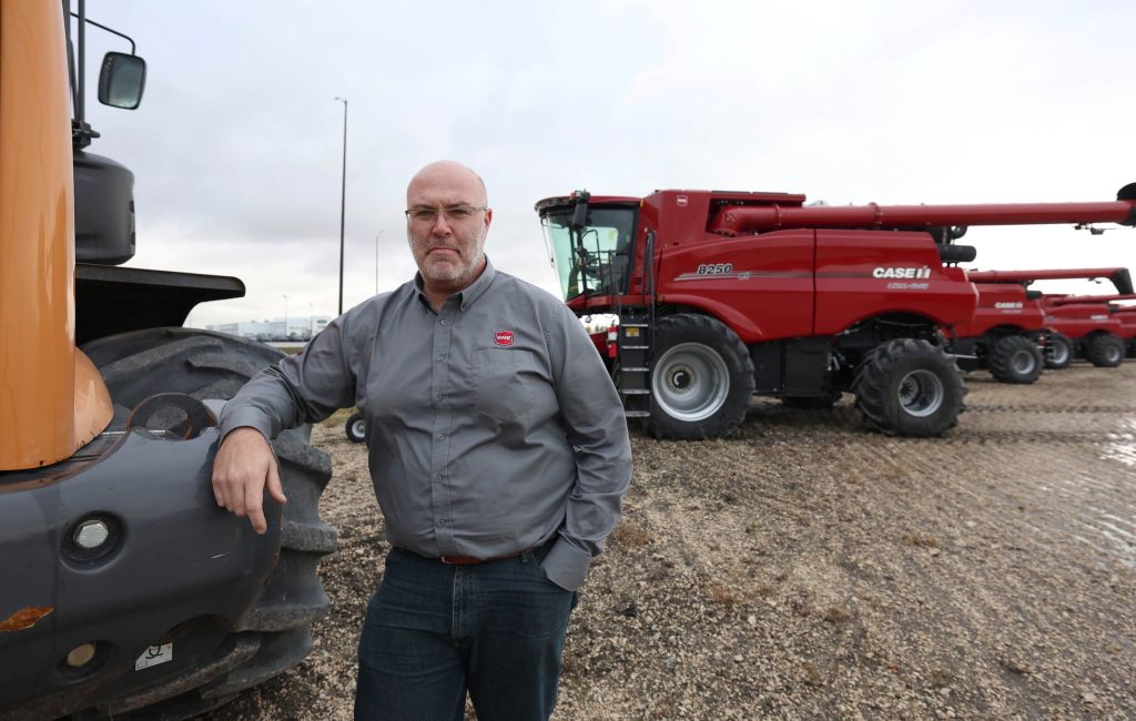 A man wearing a grey shirt leans against a tractor wheel, with anothe large tractor behind him.