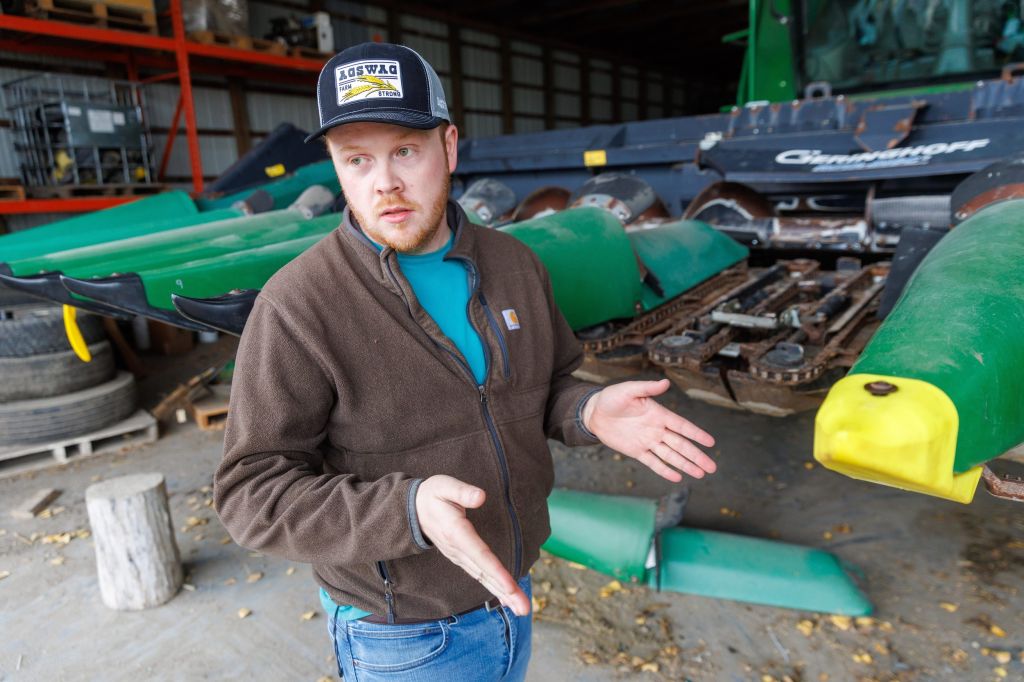 A man wearing a baseball hat and brown fleece jacket gesticulates in front of a farm storage area.
