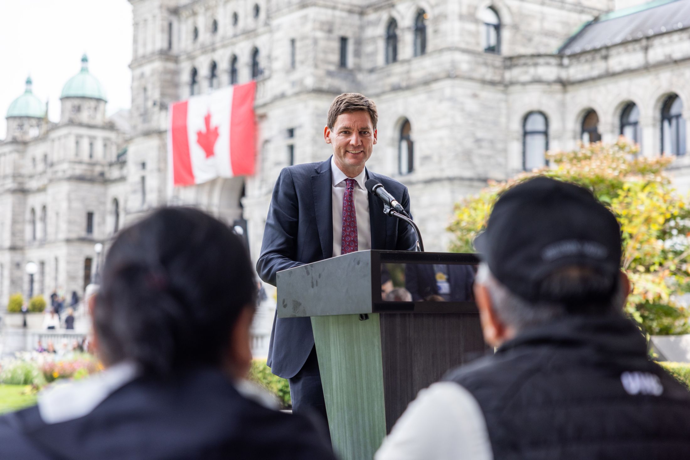 Premier David Eby stand at a podium with the BC legislature in the background. He's wearing a dark blue suit with a white shirt and red tie. There are two seated people facing him in the foreground