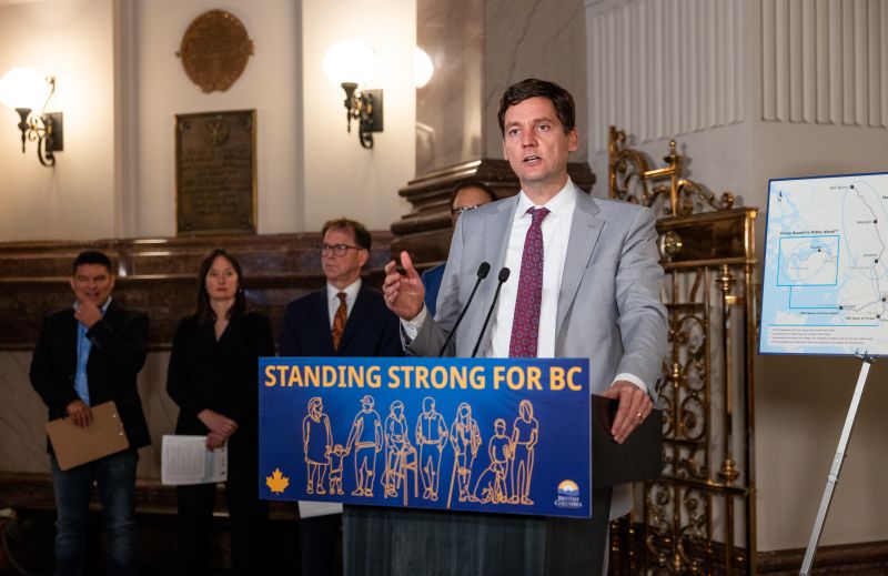 Premier David Eby stands at a "Stand Strong for BC" lectern, wearing a light grey suit, white shirt and burgundy tie