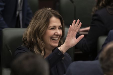 Alberta Premier Danielle Smith sits in a dark chair in the legislature, smiling and waving prior to the throne speech.