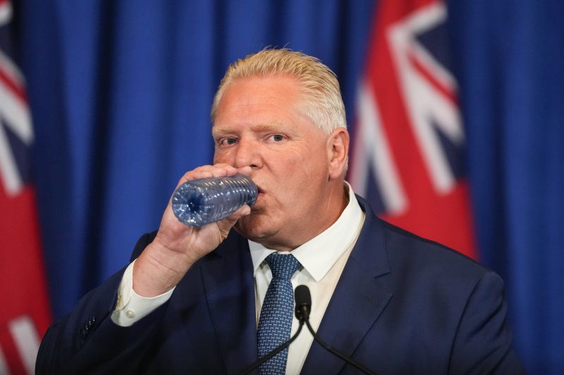 Man in a blue suit drinks from a water bottle with Ontario flag in background.