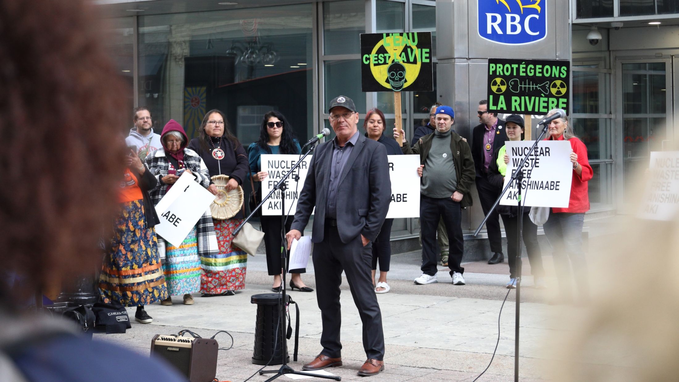 Kebaowek First Nation Chief Lance Haymond stands in front of a microphone during a rally. Behind him are people holding signs with slogans such as: “Protégeons La Rivière.”