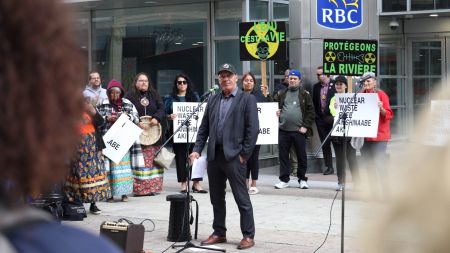 Kebaowek First Nation Chief Lance Haymond stands in front of a microphone during a rally. Behind him are people holding signs with slogans such as: “Protégeons La Rivière.”