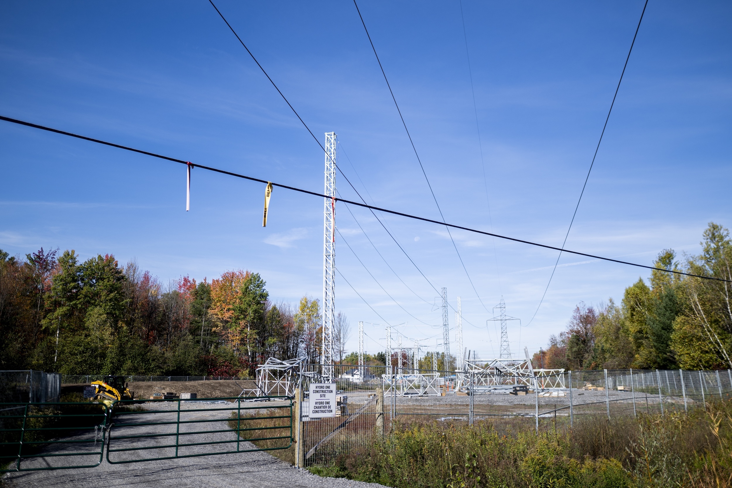 Power lines and a transmission tower behind a fence with autumn foliage on the trees behind them.