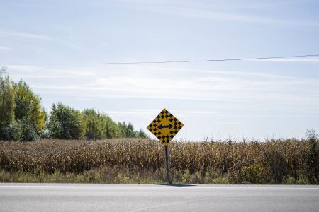 A yellow road sign with a double-arrow symbol on the edge of a road with an empty cornfield behind it.