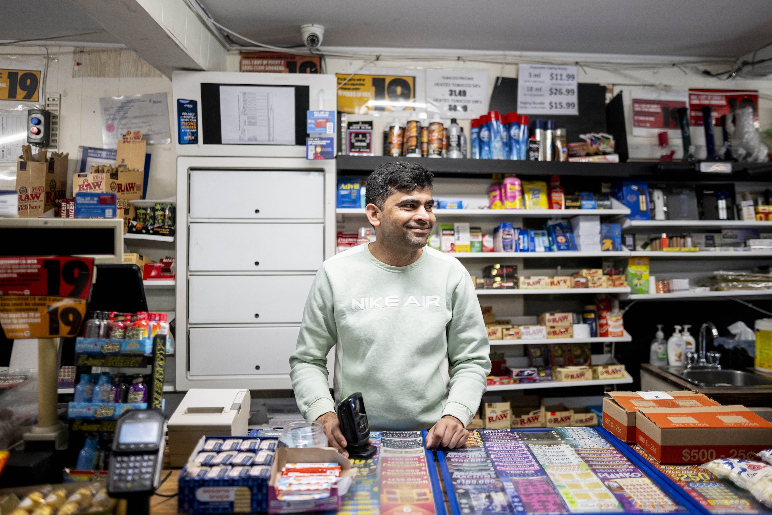 A man smiling and looking to his left, with gas station shelves filled with candies, cigarettes and other odds and ends behind him.