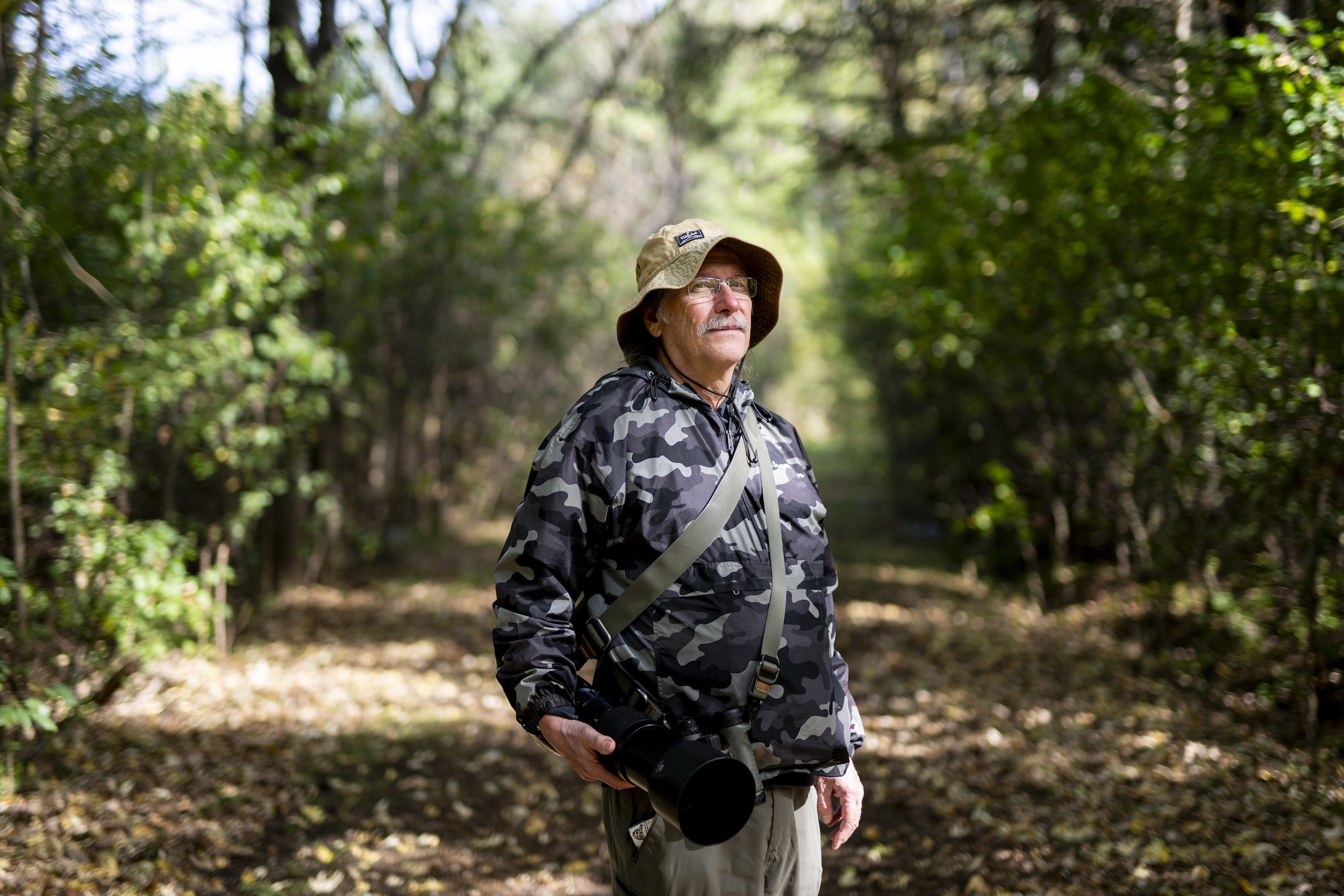 A man in a camo-printed jacket and bucket hat, carrying a large, long-lens camera, looks forward. Forested trees and a path covered in leaves fill the background.