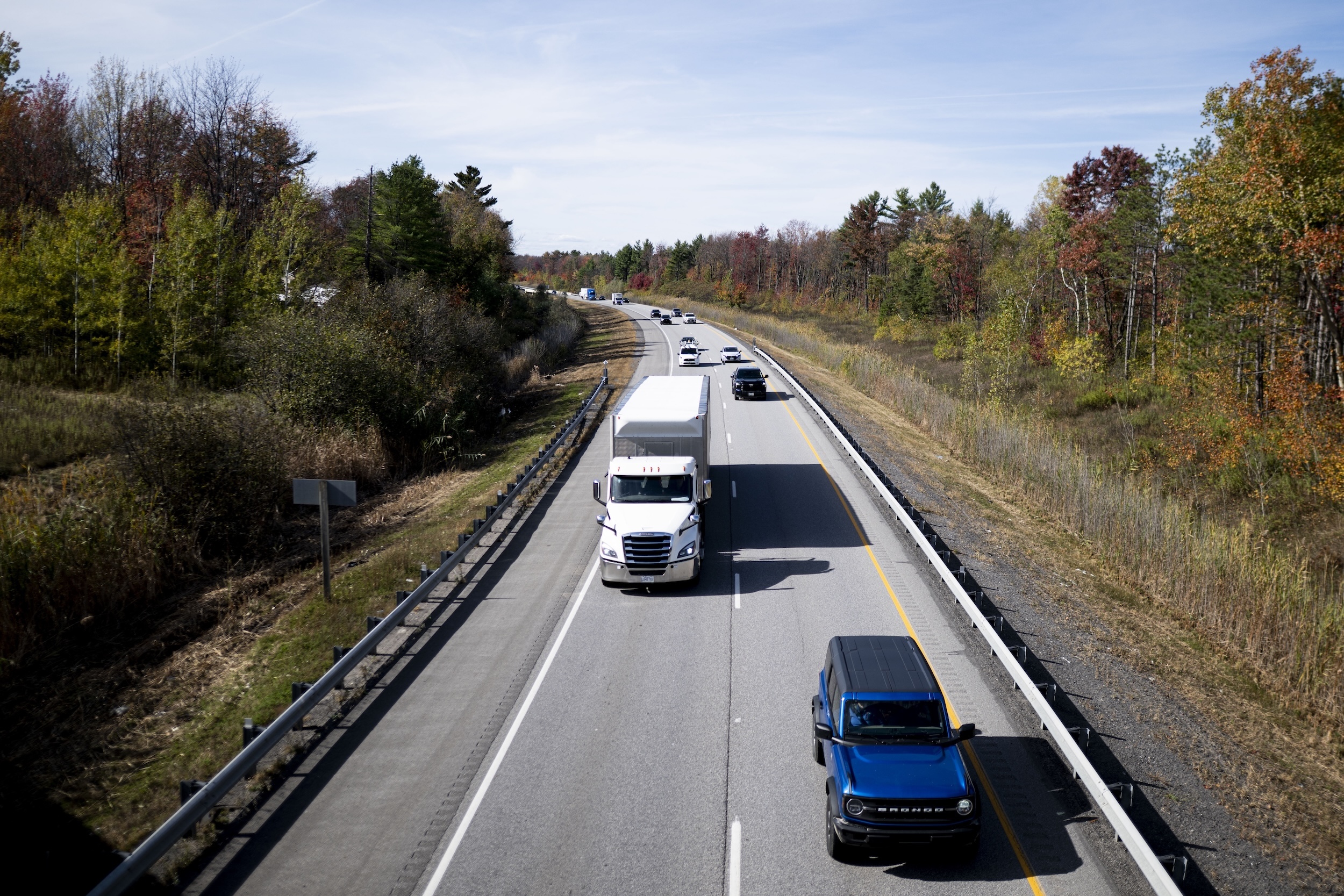 Close aerial view of several trucks barrelling down the Trans-Canada Highway, with autumn foliage on the trees on either side of the highway.