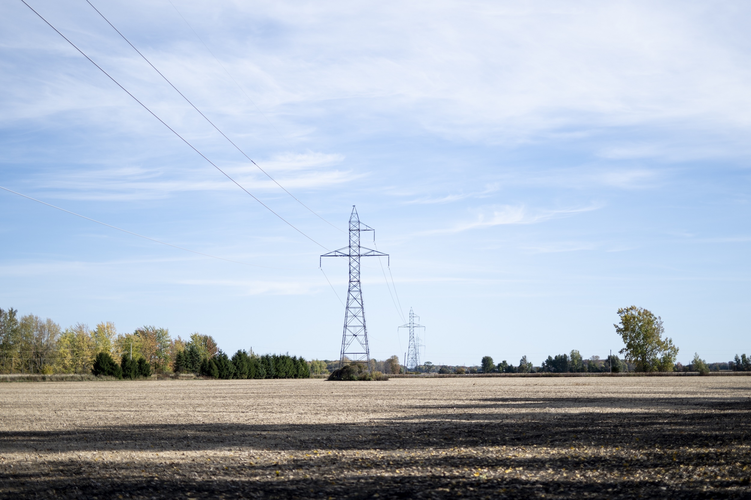 Power lines and an electrical tower in the distance in a fallow field under a cloudy sky.