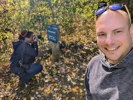 Carl Meyer takes a selfie in an autumnal forest. Behind, Spencer Colby crouches to photograph a trail sign for the Ottawa Greenbelt