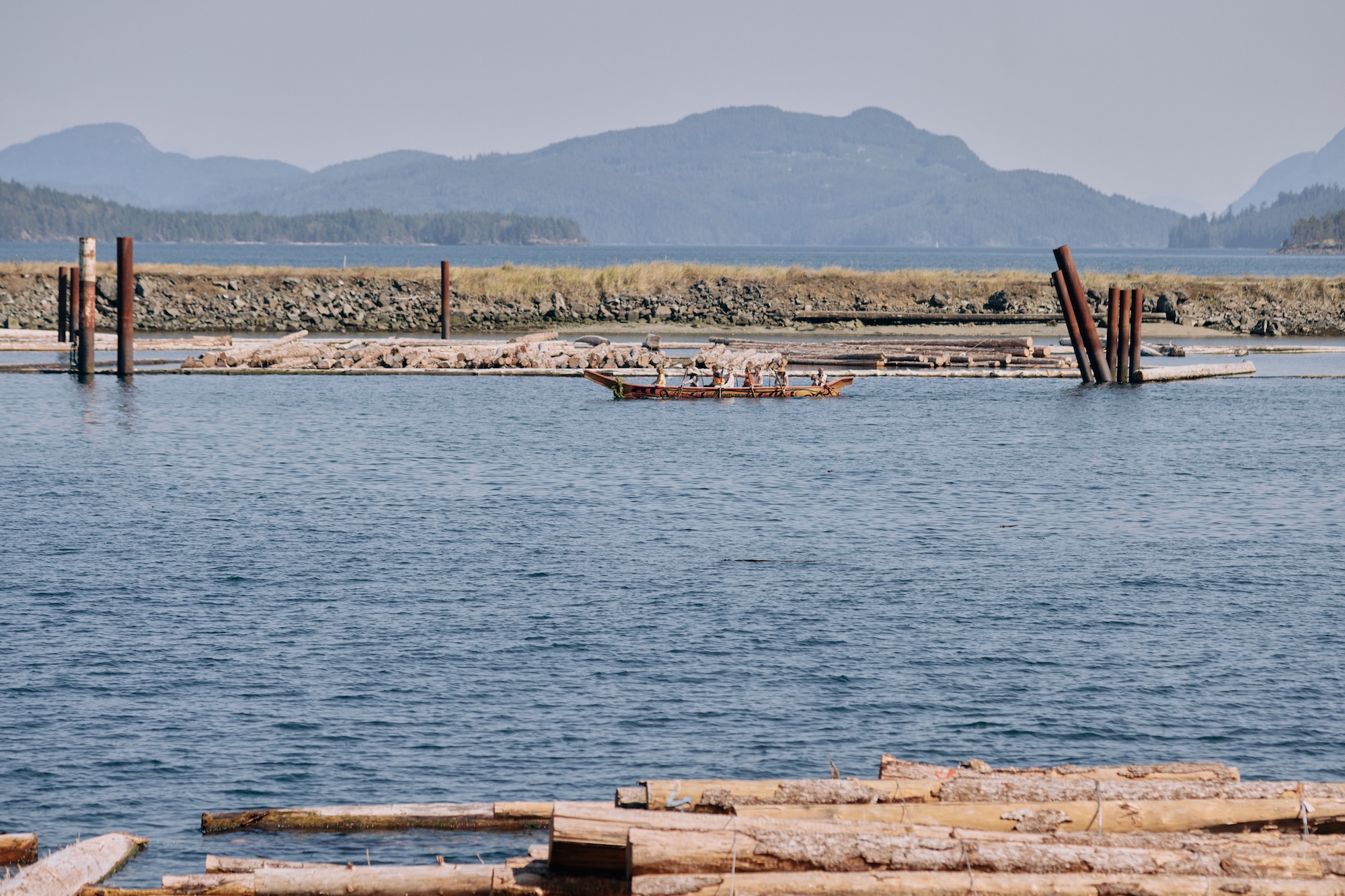 A canoe being paddled by several rowers glides through a log sort, with logs in the foregound and mountains in the background