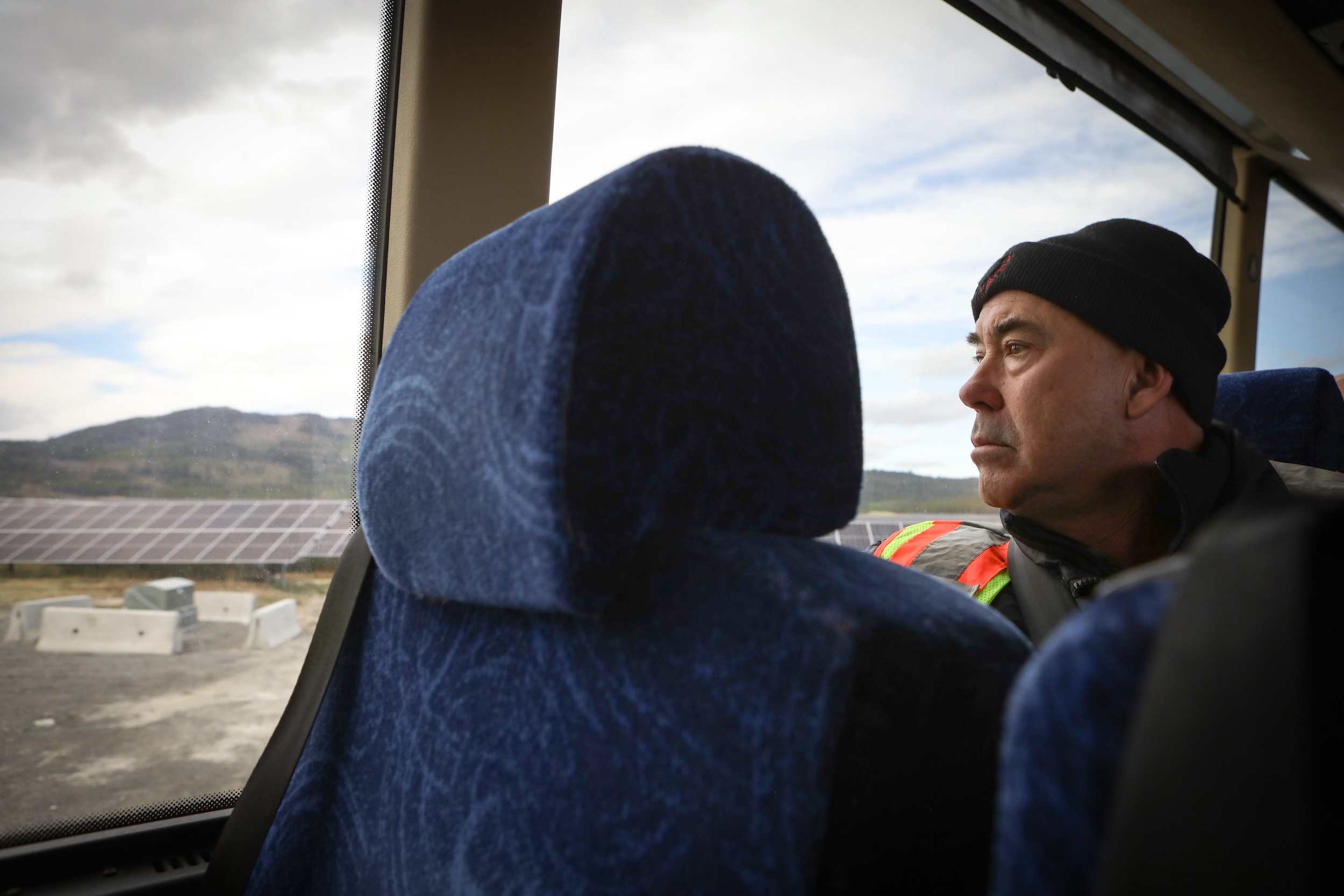 A man wearing a bright safety vest and black toque sits in a bus seat and looks out the window at a grid of solar panels.