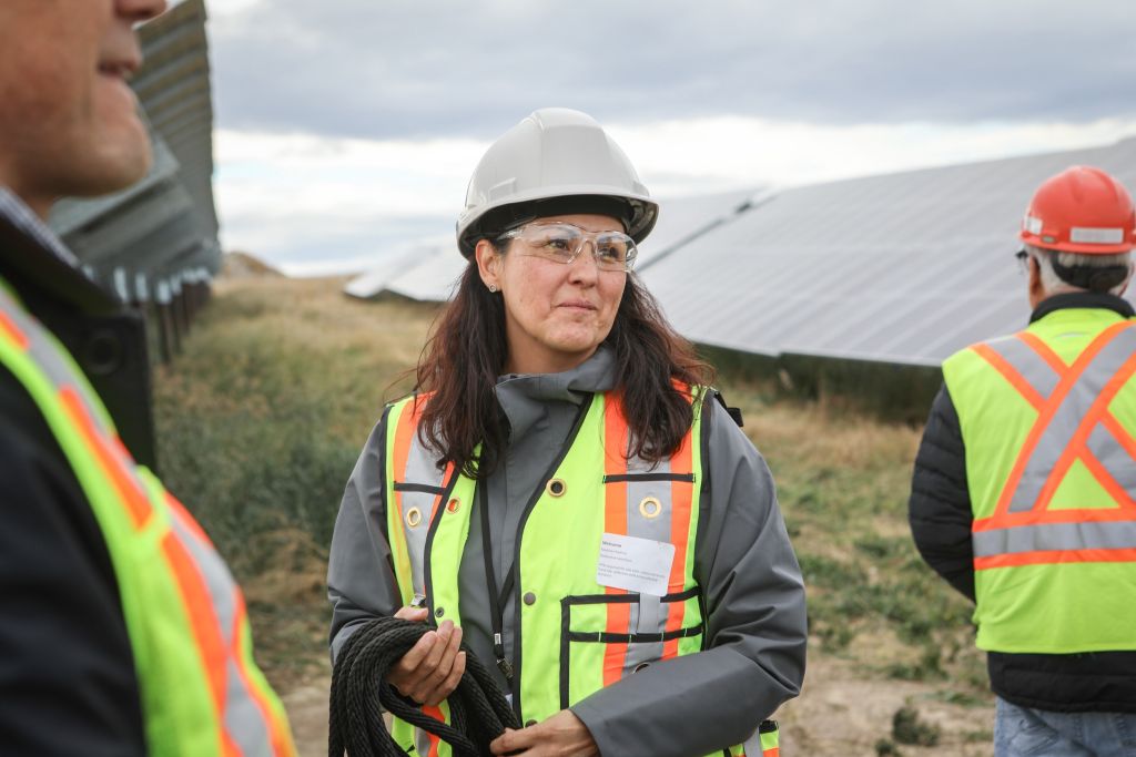 A woman wearing a bright safety vest, white hard hat and safety glasses rolls up a black cord in her hand.